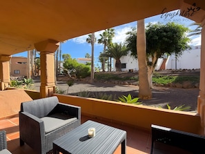 A covered outdoor seating area with wicker furniture and a table, overlooking a street with palm trees and buildings. - Oasis Dunas Eguzkilore Sun & Pool by B-Rent: Your Ideal Retreat in Corralejo (Corralejo)