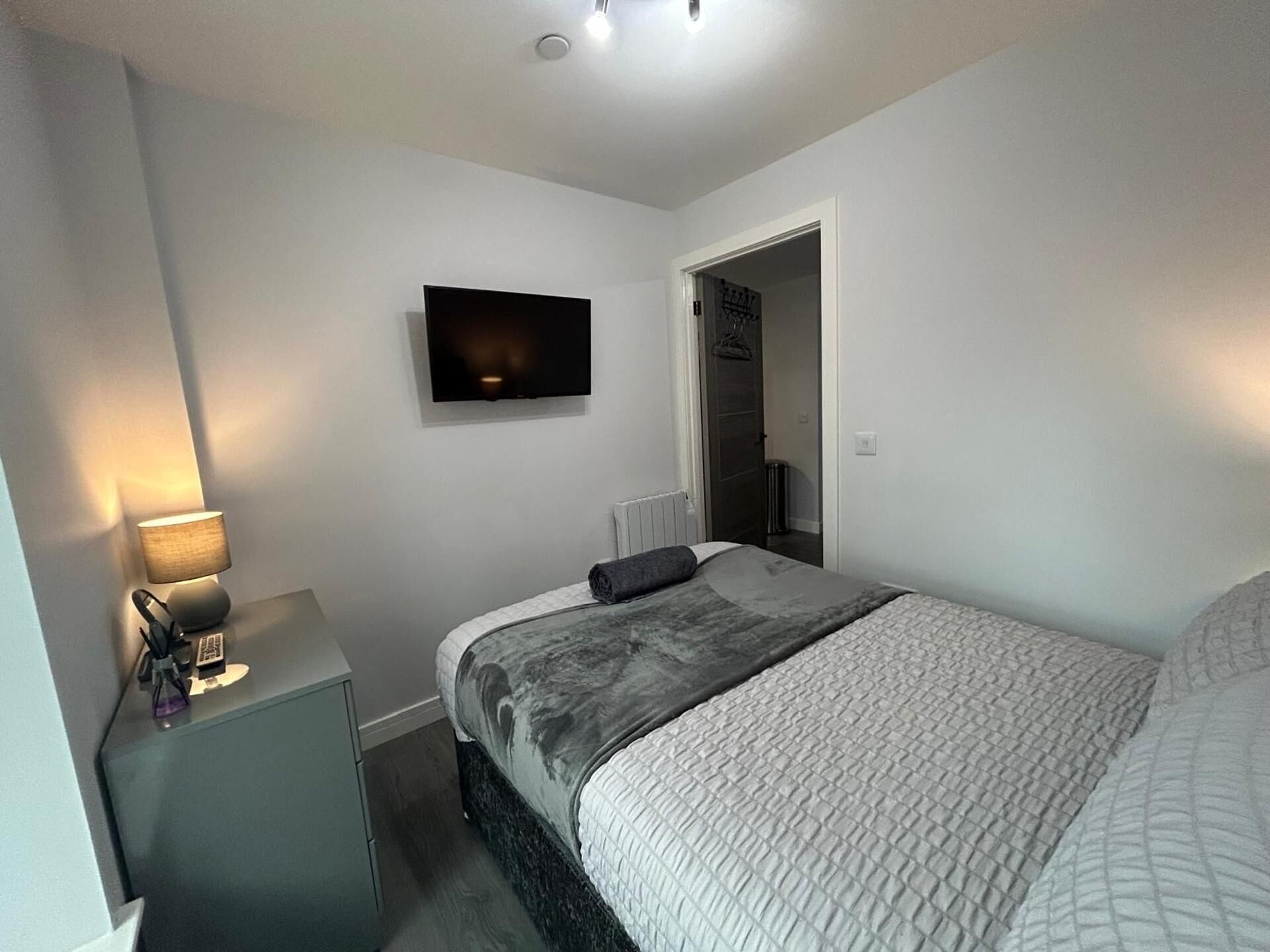 A bedroom with a bed, a TV mounted on the wall, a bedside table with a lamp, and a door leading to another room.