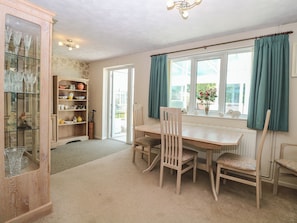 A dining area with a table and chairs, a cabinet with glassware, and a window with curtains. - 51 Colin Mclean Road (Dereham)