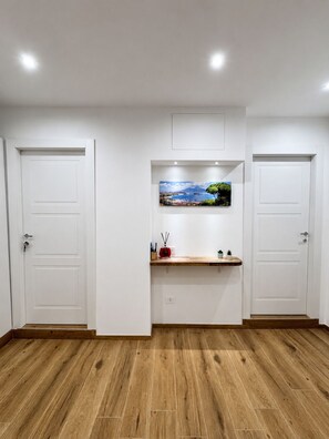 Una habitación con dos puertas blancas, suelo de madera, una estantería con un cuadro y un objeto rojo pequeño.