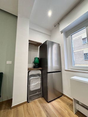 A compact kitchen with a stainless steel refrigerator, a built-in oven, and a window with a view of a brick building. - The Family Rooms (Naples)