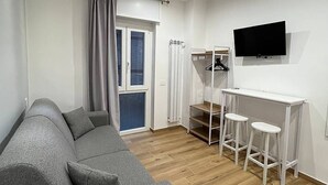 A modern living room with a grey sofa, a white desk with two stools, a flat-screen TV, and a window with curtains. - The Family Rooms (Naples)