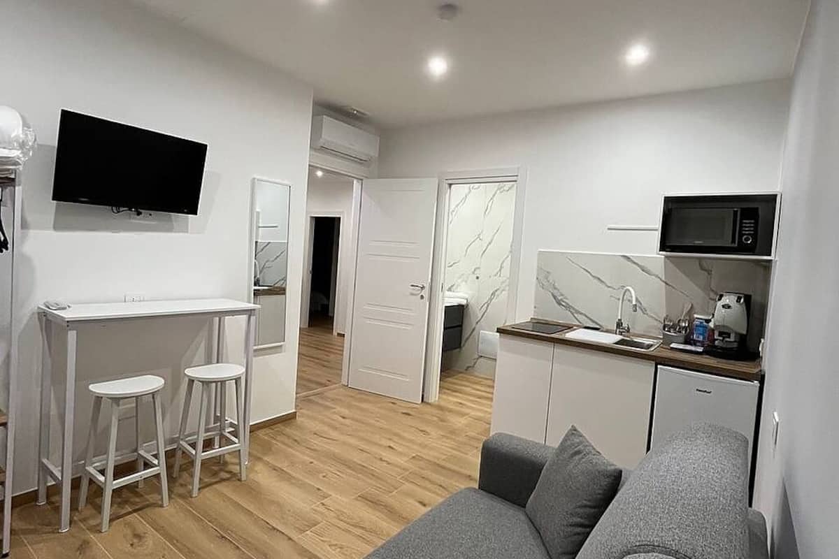 Un espacio habitable moderno y compacto, con un sofá gris, una pequeña cocina, un televisor de pantalla plana y una puerta blanca que da a otra habitación.