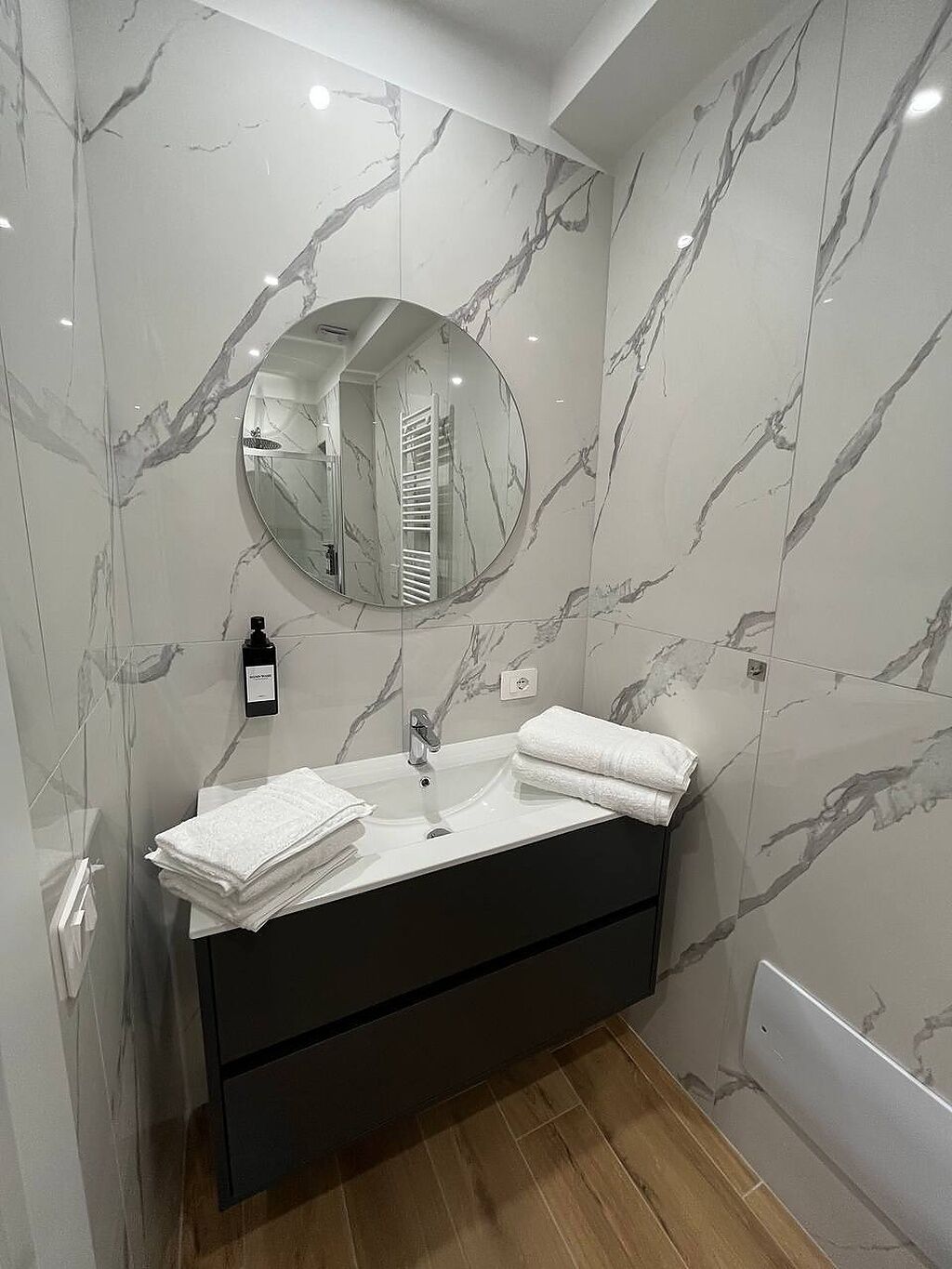 A bathroom with a marble wall, a round mirror, a sink with a black cabinet, and a towel stack.