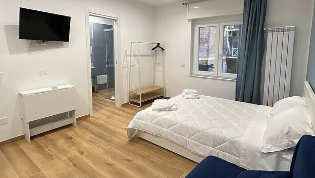 A bedroom with a bed, a TV, a desk, a window with curtains, and a radiator. - The Family Rooms (Naples)