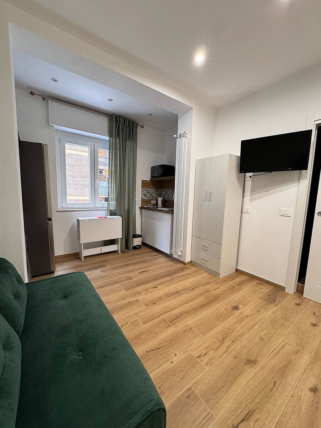 A compact room with a green sofa, a small kitchenette, a TV mounted on the wall, and a window with curtains.