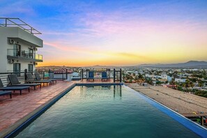 Una piscina en la azotea con sillones de descanso, vista panorámica de la ciudad y cielo despejado.