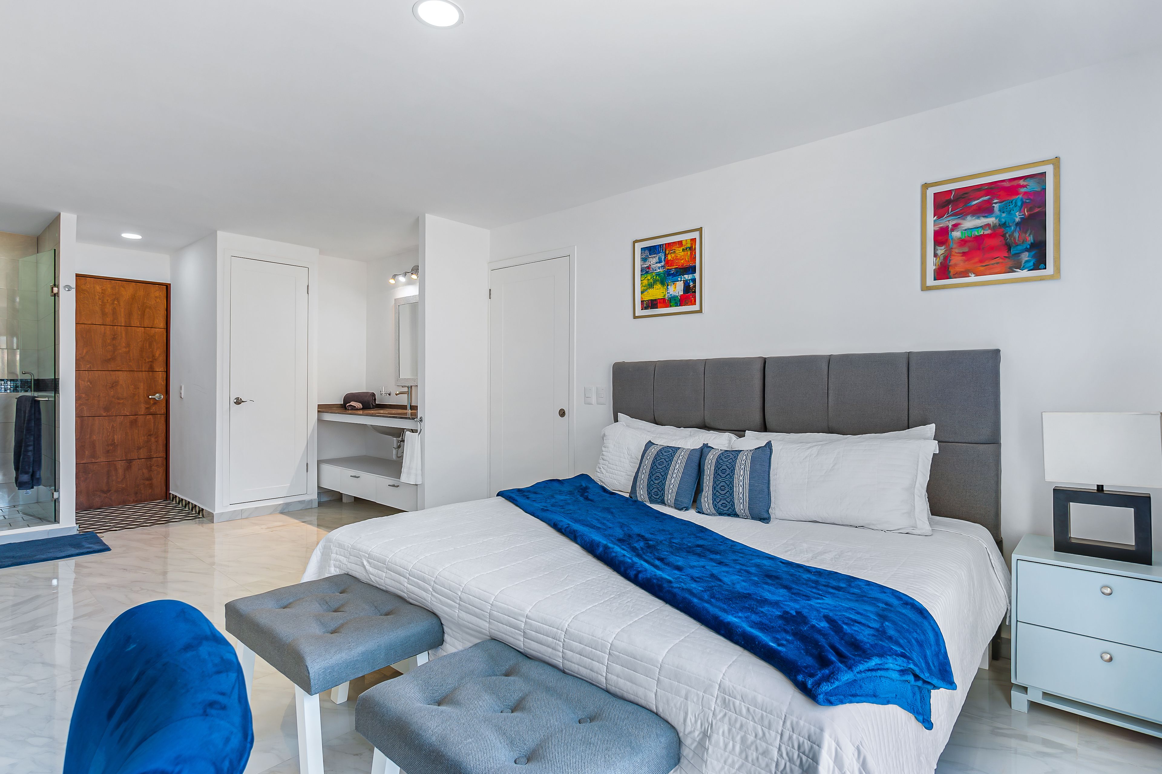 A modern bedroom with a large bed, a grey headboard, a bedside table with a lamp, and a blue bench.