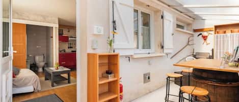 A compact room with a bed, a small table, a wooden shelf, a bar area with stools, and a window with white shutters.