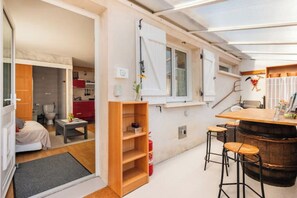 A compact room with a bed, a small table, a wooden shelf, a bar area with stools, and a window with white shutters.