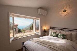 A bedroom with a bed, a bedside table with a lamp, a wall-mounted air conditioner, and a window with a view of a coastal landscape. - La finestra sul Lago by Theater Home - with marvellous lake view (Tignale)