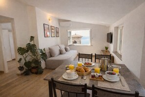 A dining area with a table set for breakfast, a sofa, and a view of a building through a window. - La finestra sul Lago by Theater Home - with marvellous lake view (Tignale)