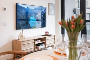 Ein modernes Wohnzimmer mit einem großen Flachbildfernseher an der Wand, einer Holzkommode mit Büchern, einer Vase mit Blumen und einem Esstisch mit einem Weinglas.