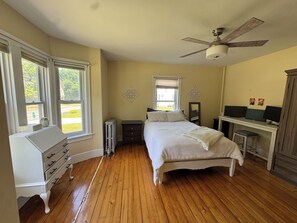 Quarto com cama, penteadeira, radiador, escrivaninha com computadores e ventoinha no teto.