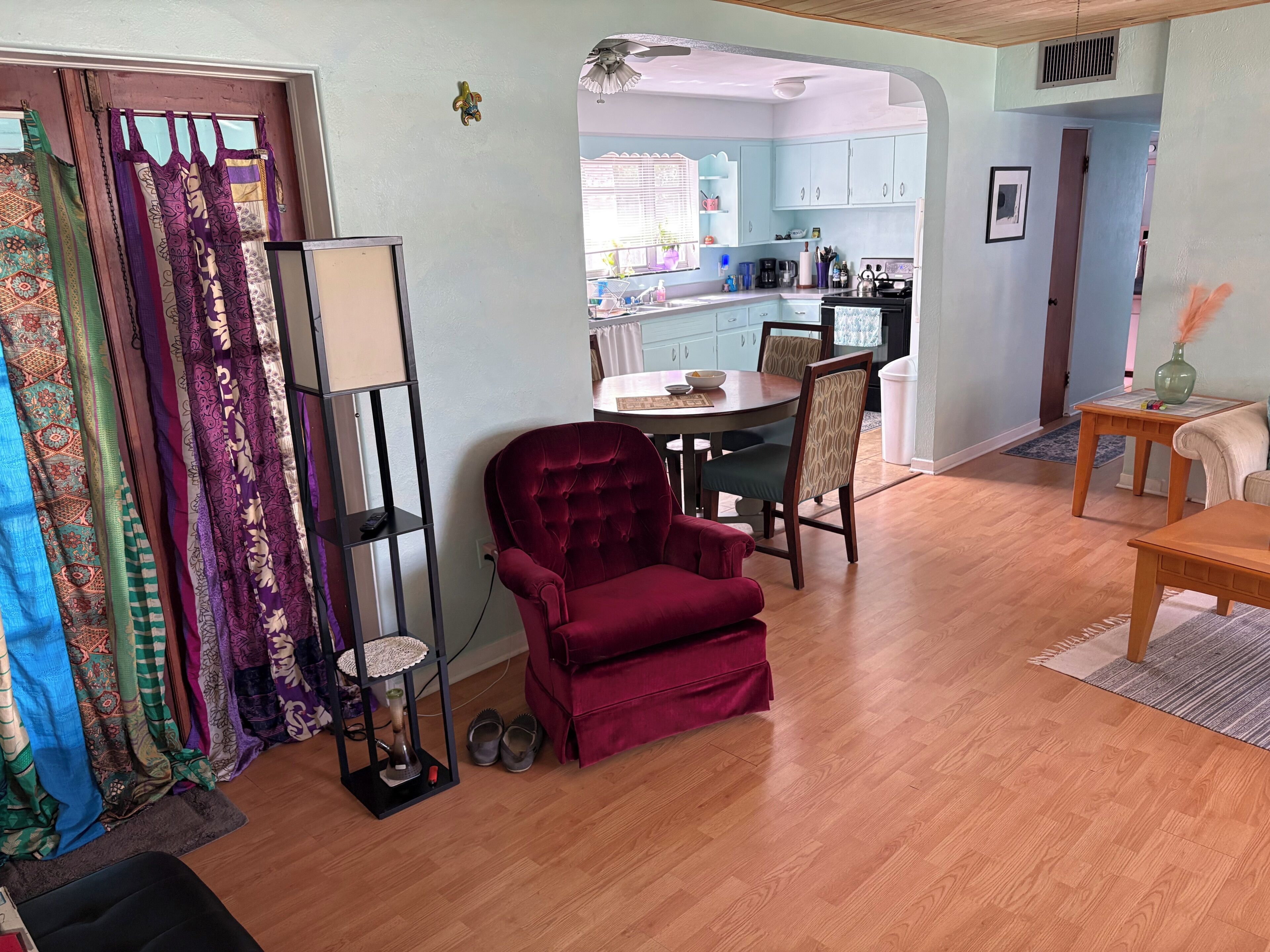 A living room with a red armchair, a coffee table, and a bookshelf. There is a kitchen visible in the background.