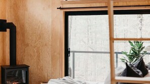 Une chambre à coucher avec un lit, un ventilateur de plafond en bois, une cheminée et de grandes fenêtres donnant sur la forêt.