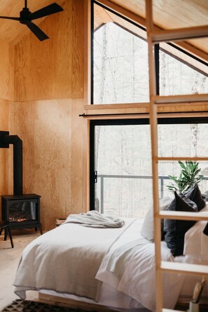 Ein Schlafzimmer mit Bett, einem Holzklimaanlage, einem Kamin und großen Fenstern mit Blick auf Bäume.