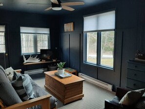 Ein Zimmer mit einer dunkel gestrichenen Wand, einem Holztisch, einer Couch, einem Schreibtisch mit Computer und großen Fenstern.