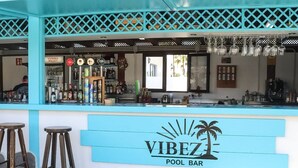 Um bar à beira da piscina com uma placa indicando 'VIBEZ POOL BAR', banquetes, um balcão com diversas garrafas e uma janela.