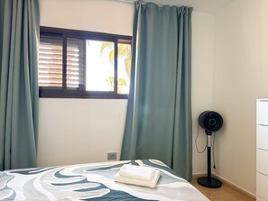 A bedroom with a bed, a window with blinds, a standing fan, and a power outlet on the wall. - Amazing Oasis Dunas Wi-Fi and Air Conditioning by B-Rent (Corralejo)