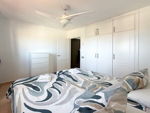 Une chambre à coucher avec un lit, un ventilateur de plafond, des armoires blanches et une porte donnant sur une autre pièce.