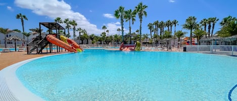 A large swimming pool with a water slide, surrounded by palm trees and lounge chairs.