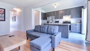 Una sala de estar moderna con un sofá seccional gris, una mesa de centro de madera y una cocina con armarios grises y electrodomésticos de acero inoxidable.
