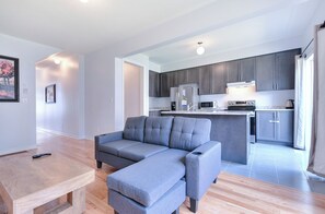 Ein modernes Wohnzimmer mit einem grauen Ecksofa, einem Holztisch und einer Küchenzeile mit grauen Schränken und Edelstahlgeräten.