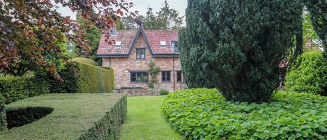 A stone house with a tiled roof, surrounded by well-maintained hedges and a neatly trimmed lawn.