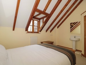 Ein Schlafzimmer mit schrägen Wänden, einem Fenster, einer Heizung, einem Nachttisch, einem Bett und einem Badezimmer mit Waschbecken und Spiegel.