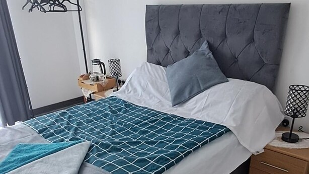 Een netjes opgemaakt bed met een grijs hoofdeinde, een nachtkastje met een lamp en een wandrek voor het drogen van was.
