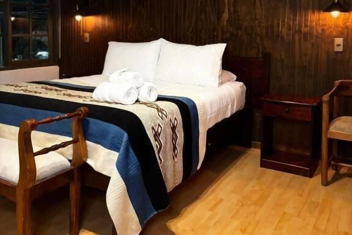A hotel room with a bed, wooden floor, a nightstand, a chair, and a large window.