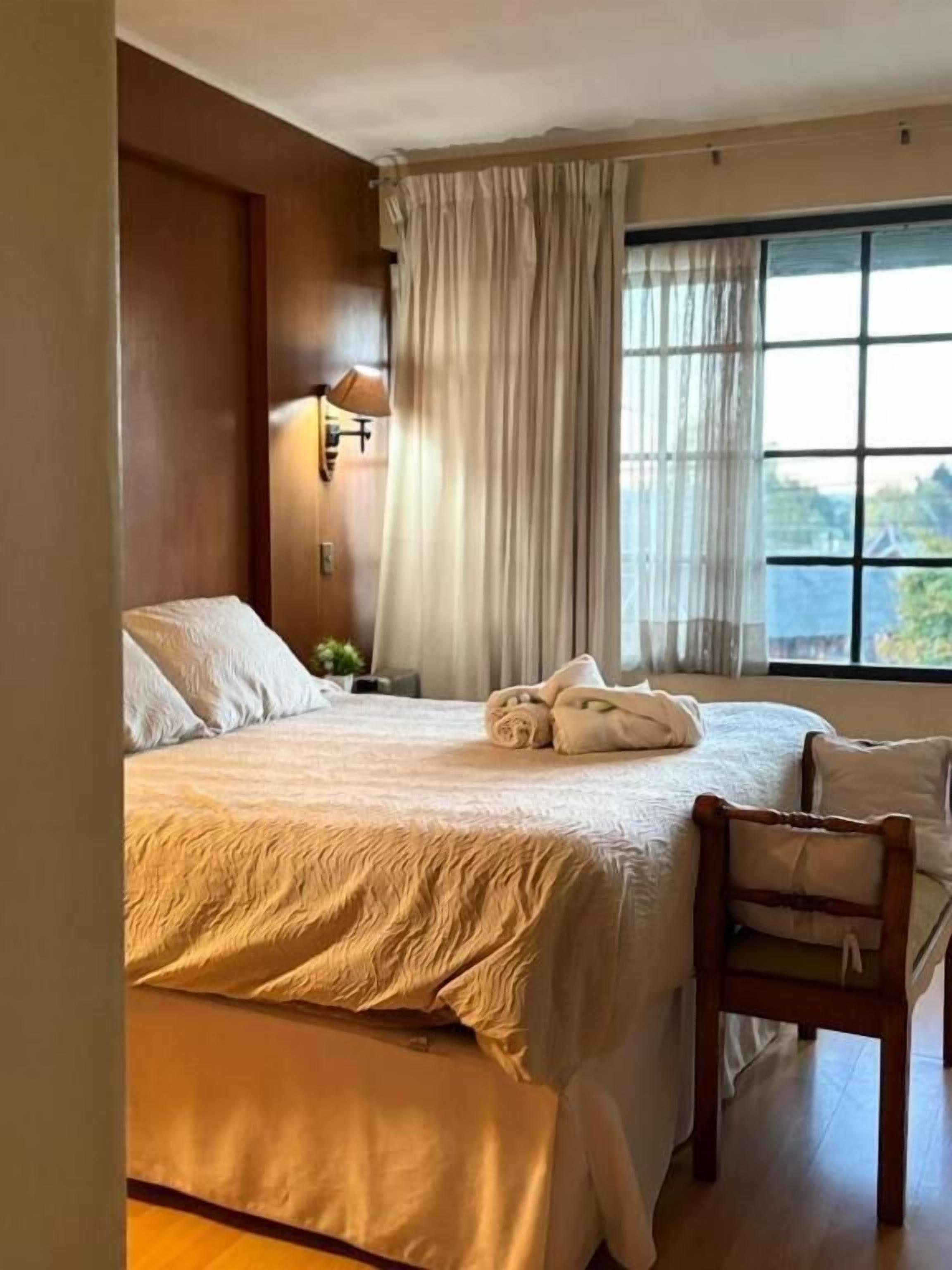 A hotel room with a bed, a chair, a window with curtains, and a lamp.
