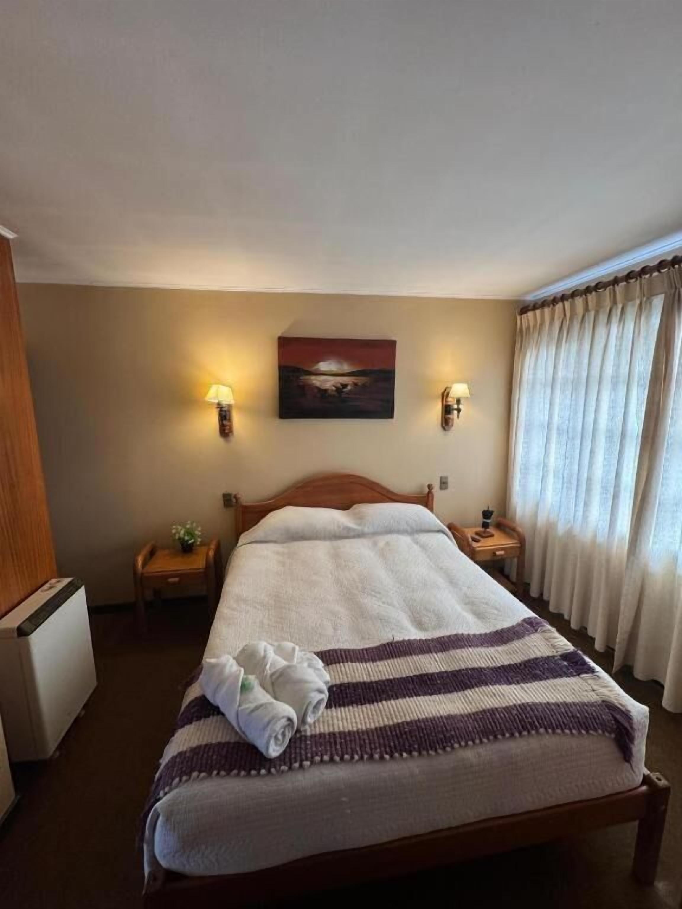 Et hotelværelse med en seng, to sengeborde med lamper, et billede på væggen og et vindue med gardiner.