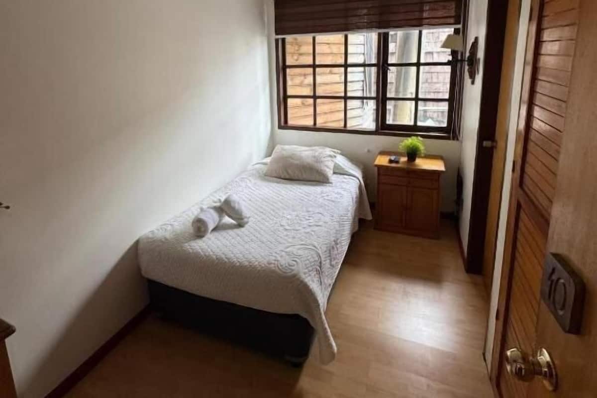 A small, clean room with a single bed, a bedside table with a lamp, and a window with blinds.