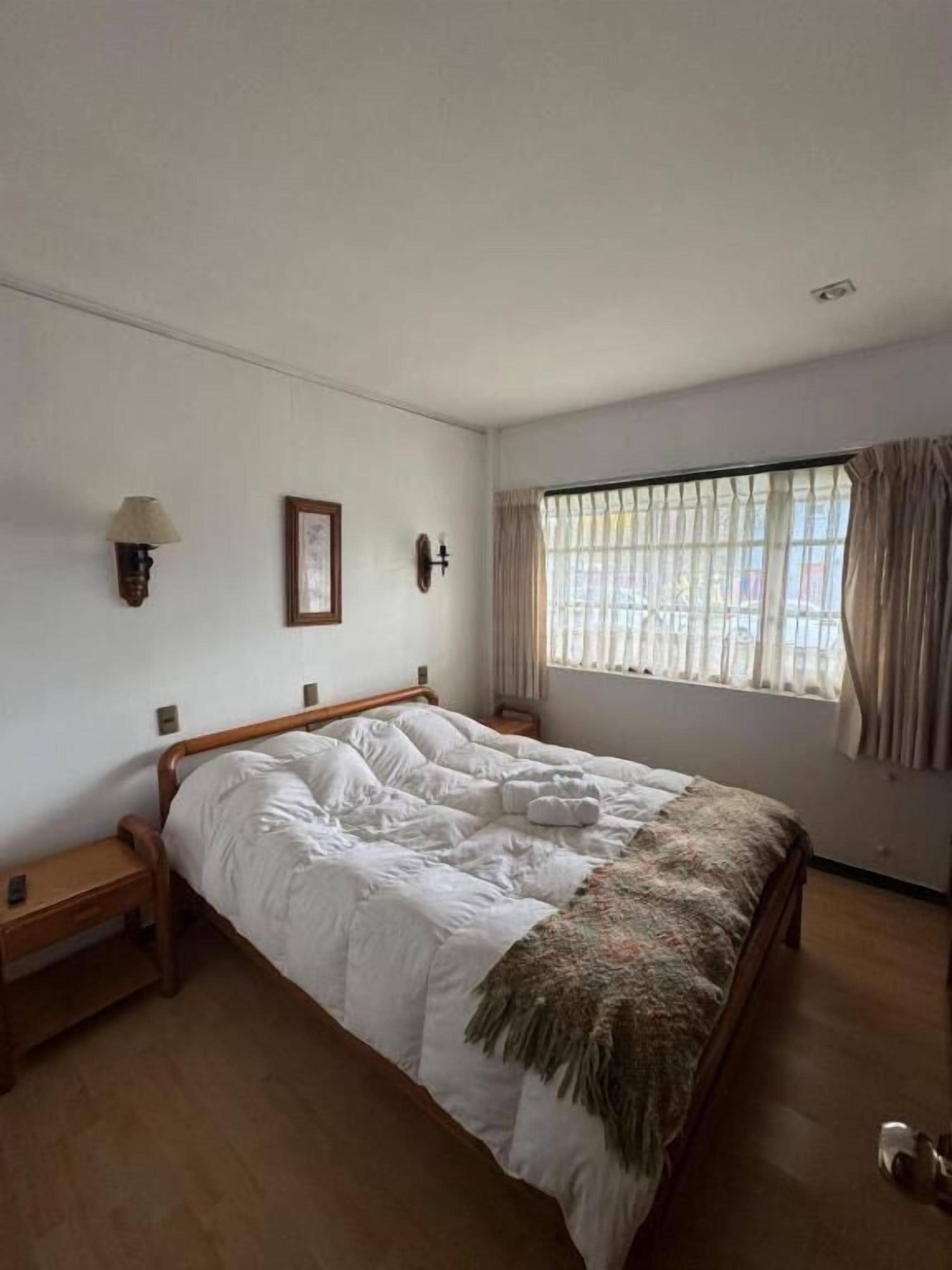 A bedroom with a bed, bedside tables, a window with curtains, and a wall-mounted lamp.