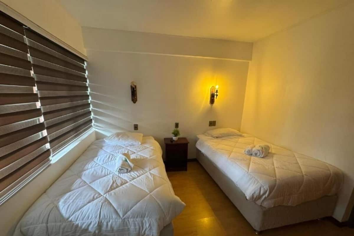 A small, clean room with two beds, a nightstand, a wall-mounted lamp, and a window with blinds.