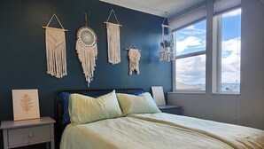 A bedroom with a bed, bedside tables, macramé wall hangings, a window with a view, and a framed picture on the nightstand. - Phinney Ridge Comfy Digs! (Seattle)