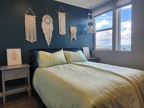 Um quarto com cama, mesas de cabeceira, painéis de macramé nas paredes, uma janela com vista e um quadro em cima da mesa de cabeceira.
