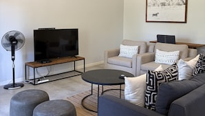 A modern living room with a flat-screen TV, two sofas, ottomans, a coffee table, and a wall-mounted fan. - Luxury 2-bedroom apartment in Victoria Falls near the falls (Victoria Falls)