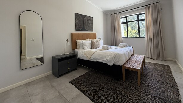 A bedroom with a bed, bedside table, lamp, ceiling fan, window with curtains, and a mirror. - Luxury 2-bedroom apartment in Victoria Falls near the falls (Victoria Falls)