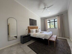 Un dormitorio con cama, mesita de noche, lámpara, ventilador de techo, ventana con cortinas y espejo.