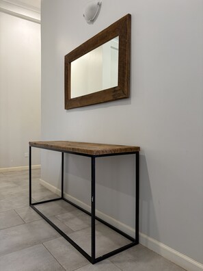 Una habitación con un espejo de marco de madera en la pared, una mesa de metal con superficie de madera y piso de baldosas.