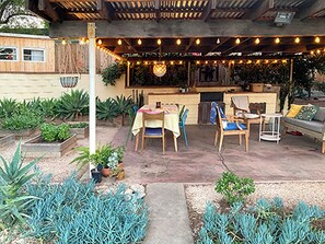 An outdoor patio with a table and chairs, a hanging light fixture, and potted plants. - Mid-century house in South Redlands with shady backyard, treehouse and pergola (Redlands)