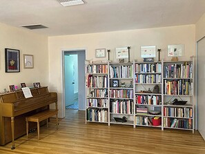Ein Zimmer mit einem Holz-Klavier, einem weißen Bücherregal voller Bücher und einer Tür zu einem anderen Raum.