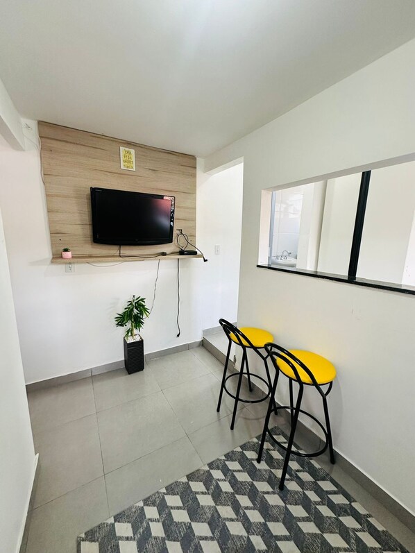 A modern room with a mounted TV, two yellow bar stools, a potted plant, and a patterned rug. - Vrbo Property (São Paulo)