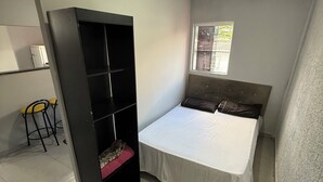 Un dormitorio con una cama, una estantería, un ventanal y un taburete pequeño.