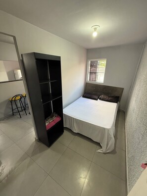 A bedroom with a bed, a bookshelf, a window, and a small stool. - Comfortable Furnished Apartment in the North Zone of São Paulo (São Paulo)