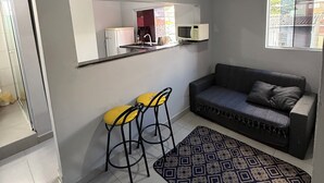 Un espacio habitable compacto con kitchenette, un sofá, dos taburetes altos y un tapete estampado.
