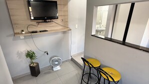 Una habitación pequeña y moderna con una televisión instalada en un estante de madera, un ventilador pequeño, dos taburetes amarillos y una alfombra estampada.